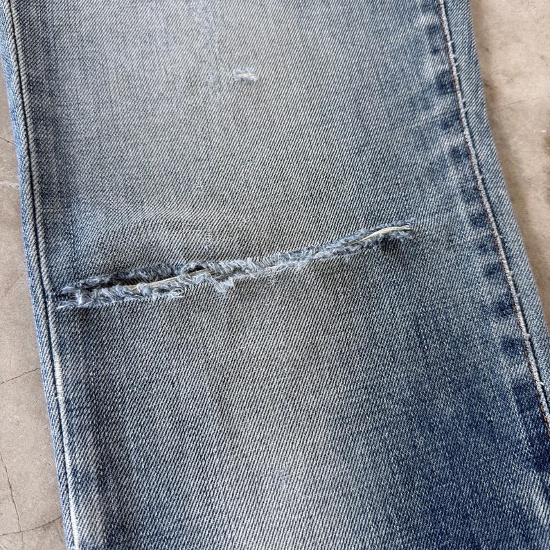May include: Close-up of light blue denim jeans with a distressed knee. The jeans show signs of wear and tear, with a large, horizontal rip across the knee area. The denim has a faded appearance.