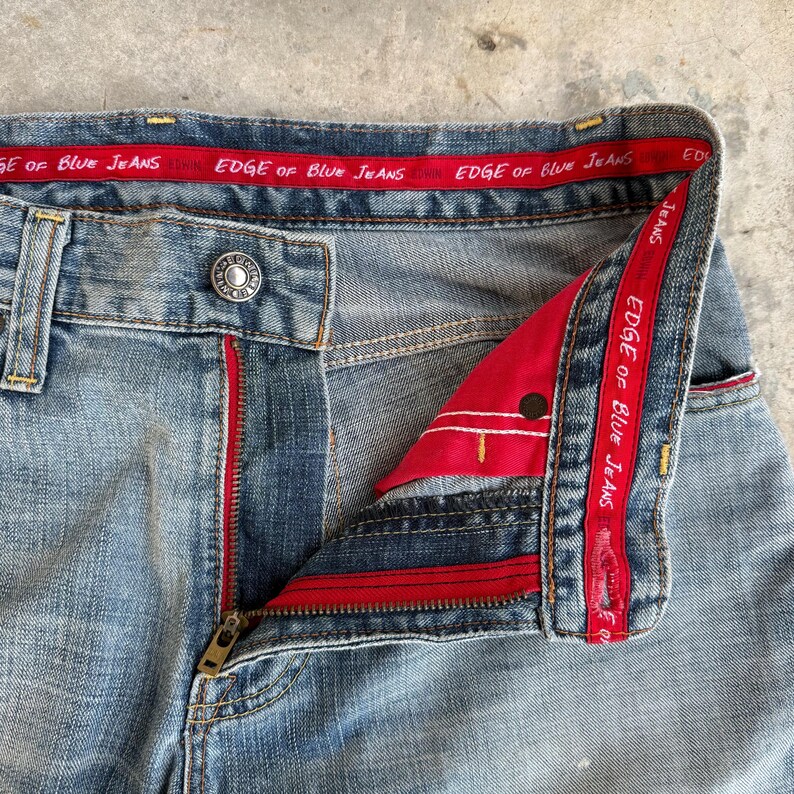 May include: Close-up of light blue denim jeans with a partially unzipped brass zipper. The jeans have a red interior lining and a red band with the text "EDGE OF BLUE JEANS". The jeans have a button closure.