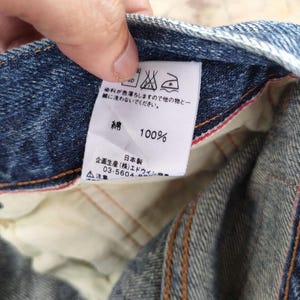 May include: Close-up of a blue denim fabric with a white tag. The tag displays Japanese text, washing instructions, and the text "100%" and "綿". The fabric has visible stitching and a red interior lining.