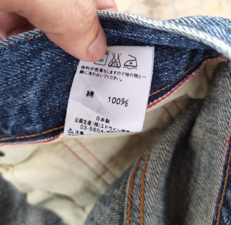 May include: Close-up of a blue denim fabric with a white tag. The tag displays Japanese text, washing instructions, and the text "100%" and "綿". The fabric has visible stitching and a red interior lining.