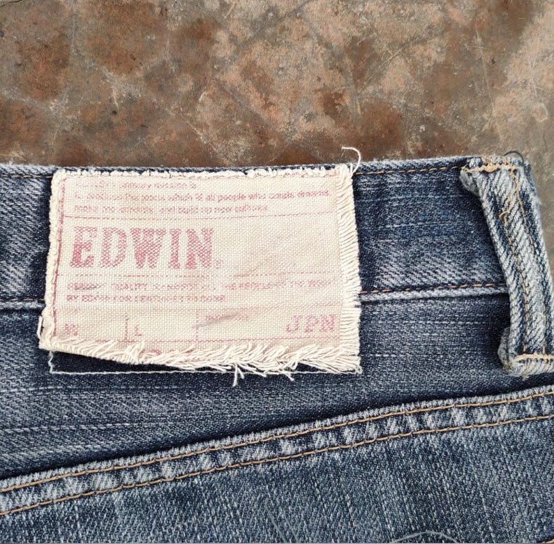 Puede incluir: Primer plano de unos vaqueros de mezclilla azules con una etiqueta rectangular de color claro. La etiqueta tiene el nombre de la marca "EDWIN" en letras rojas. Los vaqueros tienen costuras visibles y un aspecto desgastado.