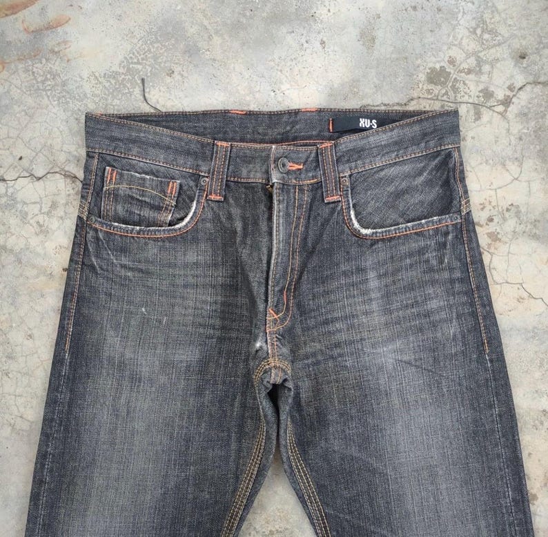 Pu&ograve; includere: Jeans in denim grigio scuro con cuciture e dettagli delle tasche arancioni. I jeans hanno un classico design a cinque tasche e una chiusura con bottoni. Il nome del marchio "XUS" &egrave; visibile in vita.