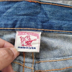 May include: Close-up of blue denim jeans featuring a True Religion label. The white label displays red text and a graphic of a Buddha playing a guitar. The text "made in USA" is also visible on the label.
