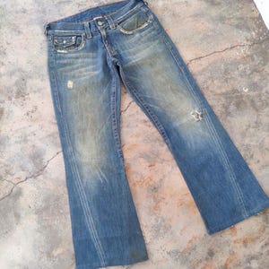 May include: Faded blue denim jeans with a distressed appearance. The jeans have a classic five-pocket design, a button and zip closure, and a flared leg. There are distressed areas on the front of the jeans.