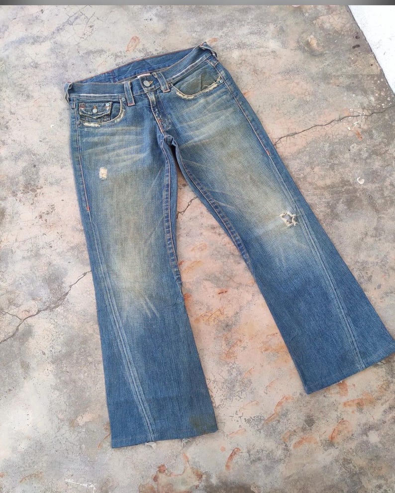 May include: Faded blue denim jeans with a distressed appearance. The jeans have a classic five-pocket design, a button and zip closure, and a flared leg. There are distressed areas on the front of the jeans.