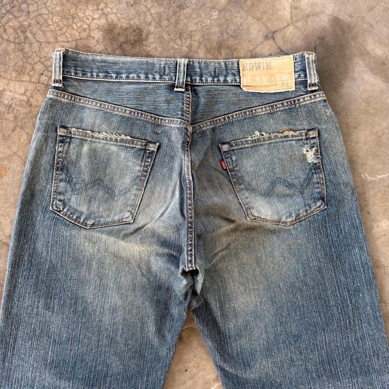 Puede incluir: Vaqueros de mezclilla azul descolorido con detalles desgastados. La vista trasera muestra los bolsillos, las trabillas y una etiqueta de cuero con la marca "EDWIN". Los vaqueros tienen un dise&ntilde;o cl&aacute;sico de cinco bolsillos y un aspecto desgastado.