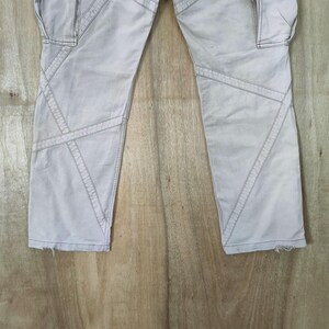 May include: Light gray cargo pants with geometric stitching details. The pants feature multiple pockets and a distressed hem. The stitching creates a unique design element, adding visual interest to the garment. The pants are made of a durable fabric.