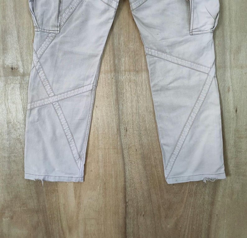 May include: Light gray cargo pants with geometric stitching details. The pants feature multiple pockets and a distressed hem. The stitching creates a unique design element, adding visual interest to the garment. The pants are made of a durable fabric.