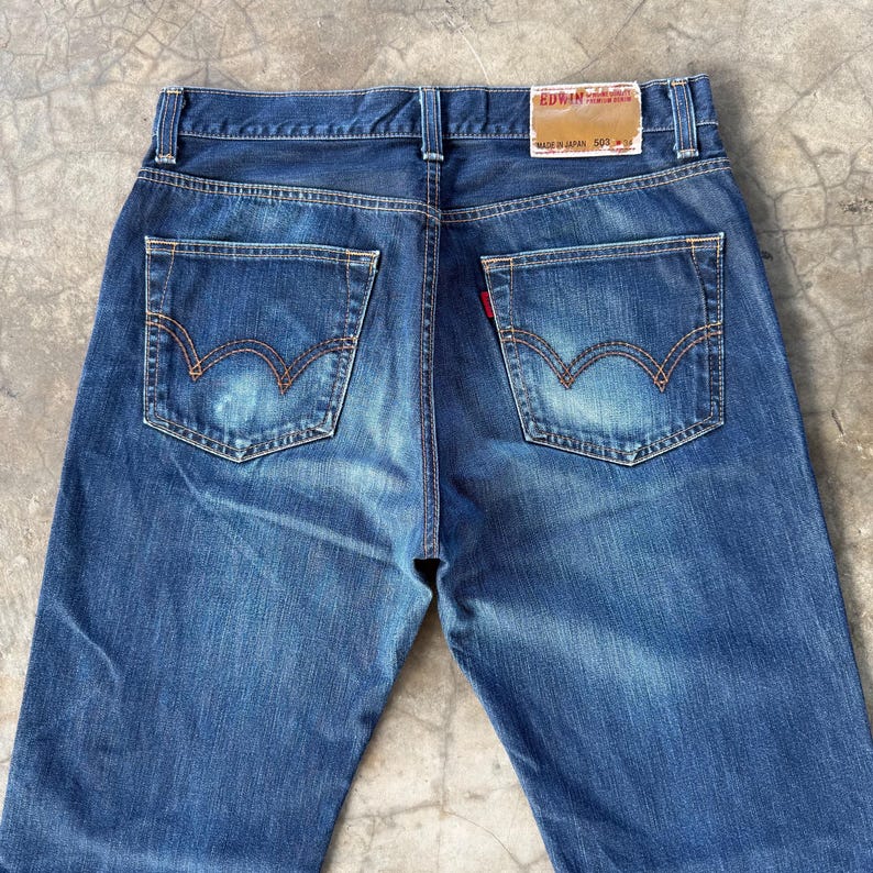 May include: Blue denim jeans with a classic five-pocket design. The jeans feature a faded wash, visible stitching, and a red tag on the back pocket. The brand name "EDWIN" is visible on a tag at the waistband.