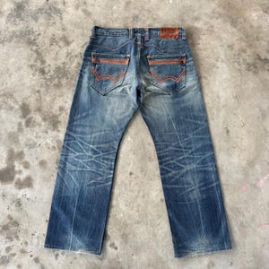 Pu&ograve; includere: Jeans in denim blu sbiadito con dettagli di cuciture arancioni sulle tasche posteriori e una toppa in pelle marrone. I jeans hanno un classico design a cinque tasche e un taglio dritto. Il marchio "EDWIN" &egrave; visibile sulla toppa in pelle.