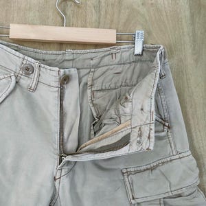 May include: Close-up of light tan cargo pants with a partially unzipped zipper and button closure. The pants have multiple pockets and are hanging on a wooden hanger against a light brown wooden background.