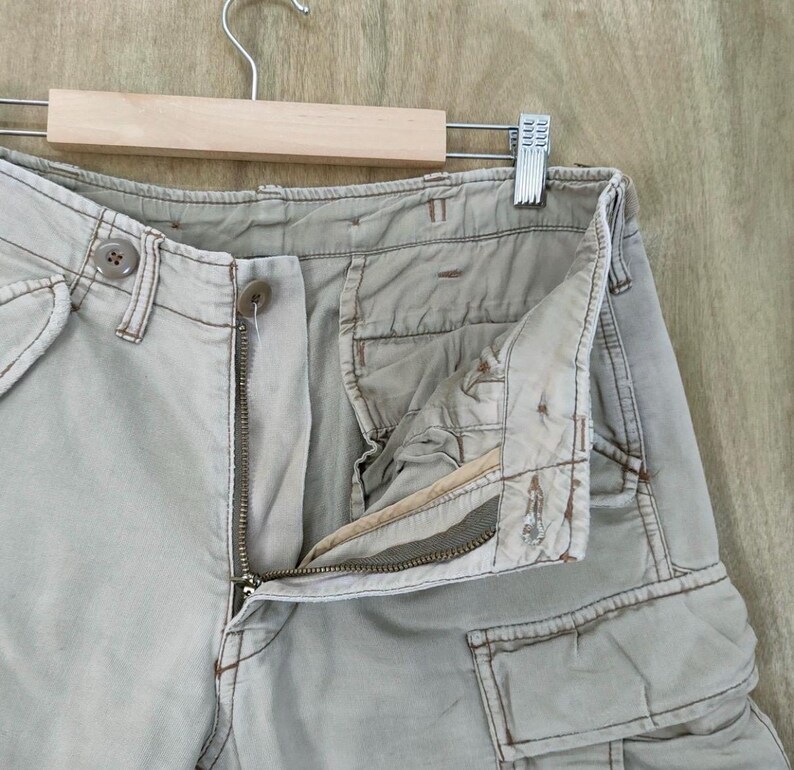 May include: Close-up of light tan cargo pants with a partially unzipped zipper and button closure. The pants have multiple pockets and are hanging on a wooden hanger against a light brown wooden background.