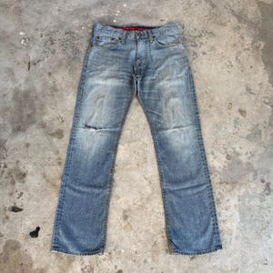 May include: Light blue denim jeans with a distressed look, featuring a ripped knee. The jeans have a classic five-pocket design, a button fly, and a red interior waistband. The jeans are laid flat on a concrete surface.