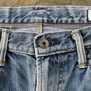 Pu&ograve; includere: Primo piano della zona della vita di jeans blu. I jeans hanno una chiusura con bottone e passanti per la cintura. Il denim &egrave; sbiadito e presenta cuciture visibili con filo giallo.