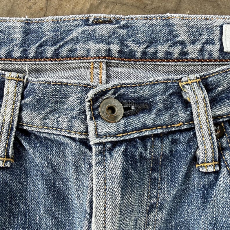 Pu&ograve; includere: Primo piano della zona della vita di jeans blu. I jeans hanno una chiusura con bottone e passanti per la cintura. Il denim &egrave; sbiadito e presenta cuciture visibili con filo giallo.