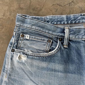 Pu&ograve; includere: Primo piano di jeans di denim blu chiaro con una tasca usurata. I jeans hanno un classico design a cinque tasche, con usura visibile. Una piccola etichetta bianca con la lettera "E" &egrave; visibile sulla tasca.