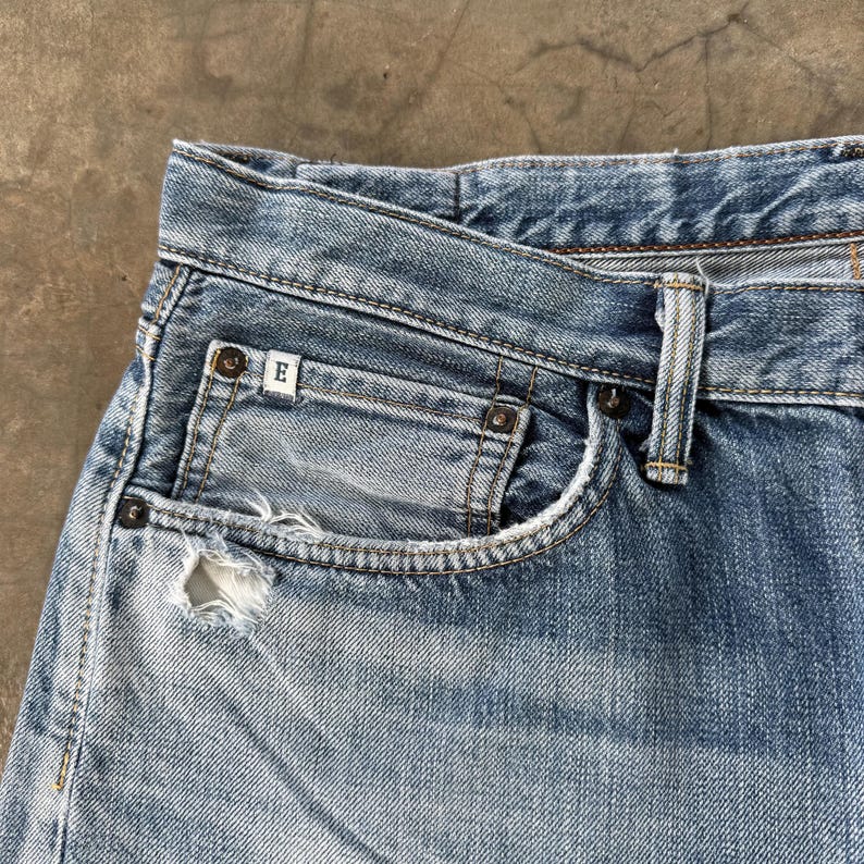 Pu&ograve; includere: Primo piano di jeans di denim blu chiaro con una tasca usurata. I jeans hanno un classico design a cinque tasche, con usura visibile. Una piccola etichetta bianca con la lettera "E" &egrave; visibile sulla tasca.