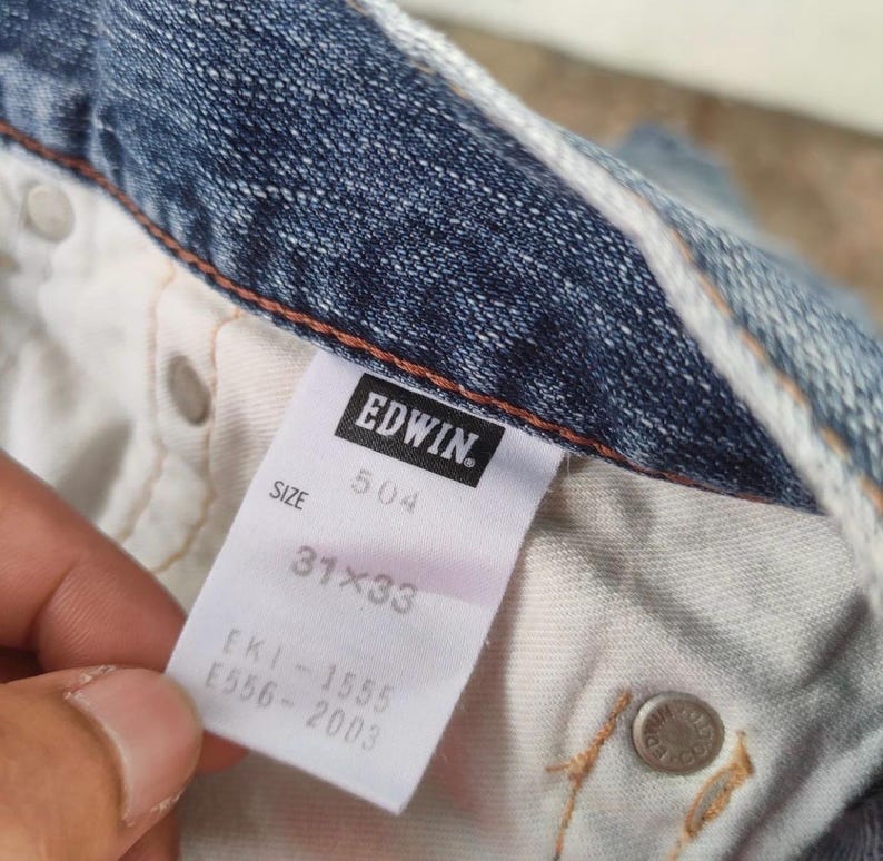 May include: Close-up of a pair of blue denim jeans. The interior label reads "EDWIN" and displays the size as 31x33. The jeans have a classic five-pocket design with metal rivets and a button closure.