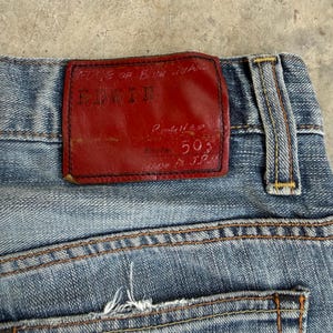 May include: Close-up of light blue denim jeans with a red leather patch. The patch features the brand name "EDWIN" and the text "EDGE OF BLUE JEANS". The jeans have visible stitching and a back pocket.