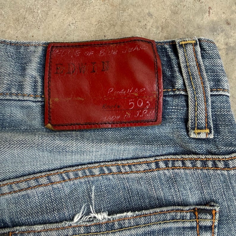 May include: Close-up of light blue denim jeans with a red leather patch. The patch features the brand name "EDWIN" and the text "EDGE OF BLUE JEANS". The jeans have visible stitching and a back pocket.