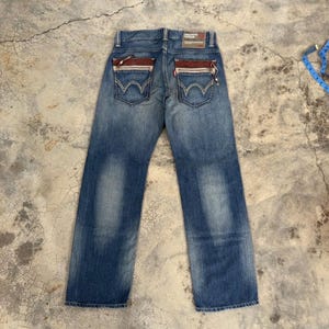 May include: Blue denim jeans with a faded wash. The back pockets have a unique design with red and white accents. The jeans have a straight leg cut and a brown leather patch.