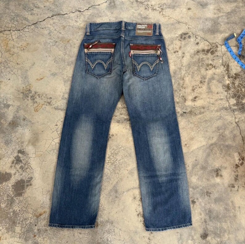 May include: Blue denim jeans with a faded wash. The back pockets have a unique design with red and white accents. The jeans have a straight leg cut and a brown leather patch.
