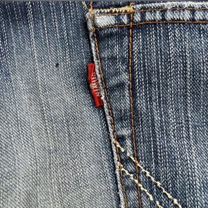 May include: Close-up of blue denim jeans with a small red tag on the back pocket. The tag has white text. The jeans have visible stitching in a contrasting brown color. The denim has a faded, worn appearance.