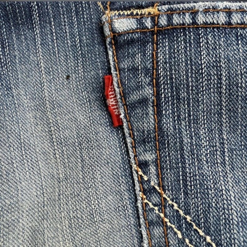 May include: Close-up of blue denim jeans with a small red tag on the back pocket. The tag has white text. The jeans have visible stitching in a contrasting brown color. The denim has a faded, worn appearance.