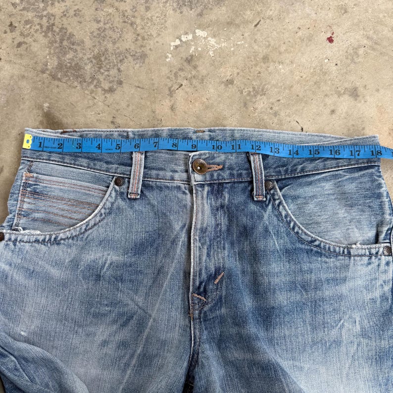 May include: Light blue denim jeans with a faded wash and visible wear. A measuring tape shows the waist size. The jeans have classic five-pocket styling, a button closure, and a zipper fly. The jeans are vintage.