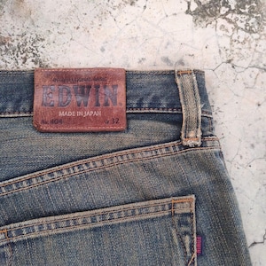 May include: Close-up of blue denim jeans with a brown leather patch. The patch reads "EDWIN" and "MADE IN JAPAN". The jeans are size W32 and have a faded appearance.