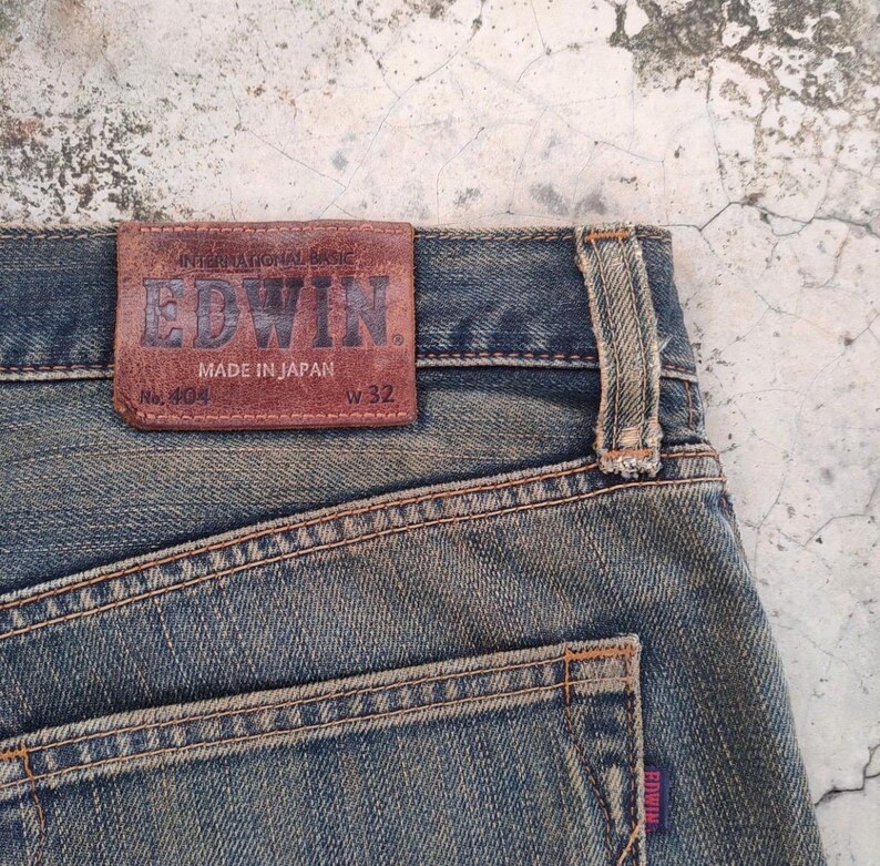 May include: Close-up of blue denim jeans with a brown leather patch. The patch reads "EDWIN" and "MADE IN JAPAN". The jeans are size W32 and have a faded appearance.