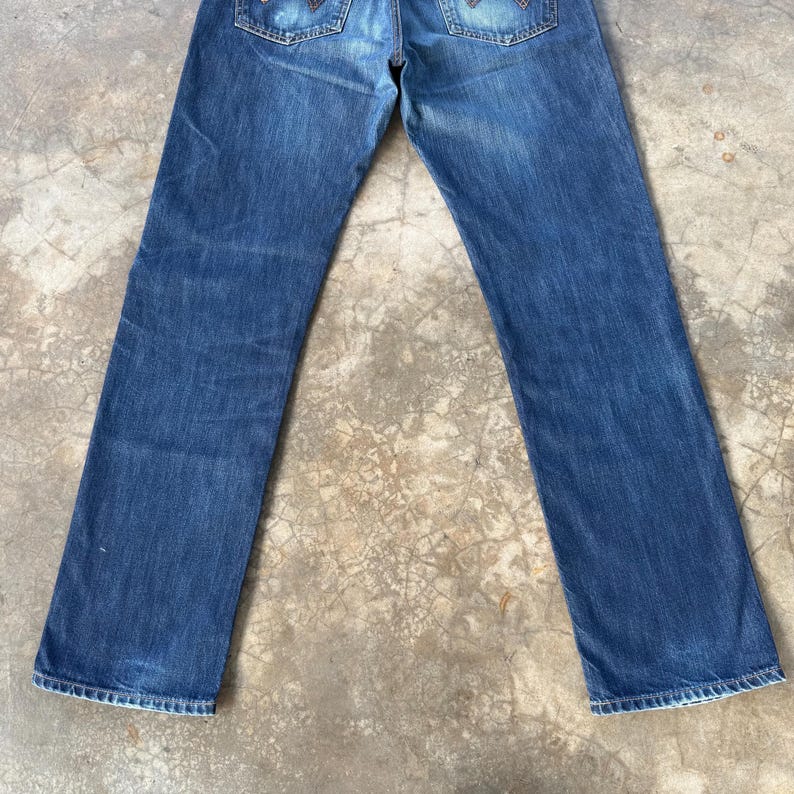 May include: Blue denim jeans with a classic five-pocket design. The jeans have a faded wash and a straight leg cut. The back pockets feature decorative stitching. The jeans are laid flat on a concrete surface.