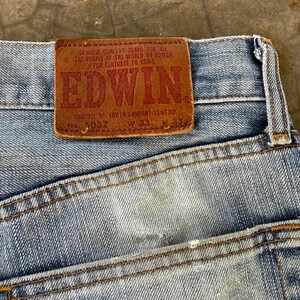 May include: Close-up of light blue denim jeans with a brown leather patch. The patch features the brand name "EDWIN" in large red letters, along with text that reads "GENUINE QUALITY JEANS FOR ALL THE PEOPLE OF THE WORLD BY EDWIN FOR CENTURIE TO COME".