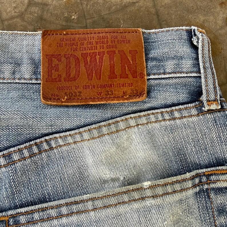 May include: Close-up of light blue denim jeans with a brown leather patch. The patch features the brand name "EDWIN" in large red letters, along with text that reads "GENUINE QUALITY JEANS FOR ALL THE PEOPLE OF THE WORLD BY EDWIN FOR CENTURIE TO COME".