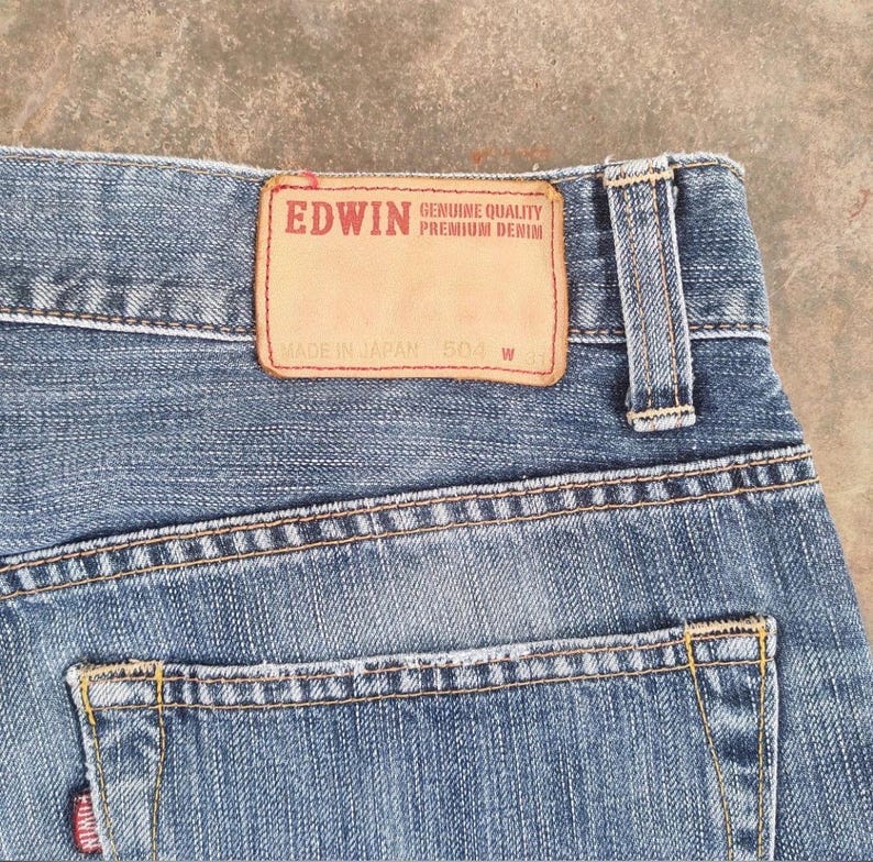 May include: Close-up of blue denim jeans with a leather patch that reads "EDWIN GENUINE QUALITY PREMIUM DENIM". The jeans have a back pocket and a belt loop. The tag reads "MADE IN JAPAN 504 W 31".