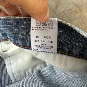 May include: Close-up of a blue denim garment's interior, showcasing a white tag with Japanese text and symbols. The tag indicates the fabric composition is 100% cotton and features a leather label. The garment's stitching and fabric texture are visible.