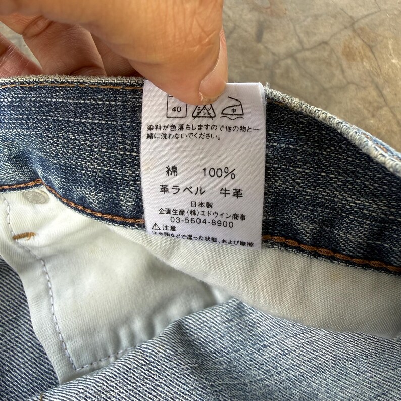May include: Close-up of a blue denim garment's interior, showcasing a white tag with Japanese text and symbols. The tag indicates the fabric composition is 100% cotton and features a leather label. The garment's stitching and fabric texture are visible.