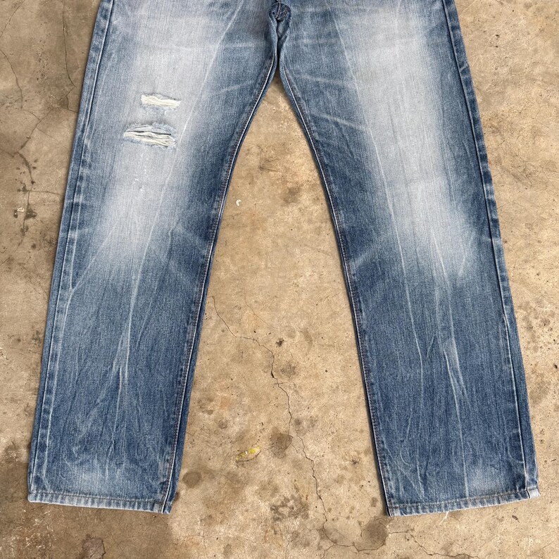 May include: Faded blue denim jeans with distressed details. The jeans have a classic five-pocket design, a button and zipper closure, and a relaxed fit. The denim has a worn appearance with light fading and a distressed area on the left knee.