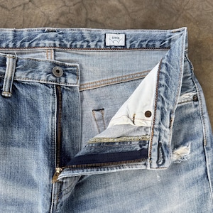 Pu&ograve; includere: Primo piano di jeans di denim blu chiaro con cerniera parzialmente aperta. I jeans hanno una chiusura con bottone e una piccola etichetta con il marchio "EDWIN". Il denim ha un aspetto sbiadito e usurato con alcuni dettagli strappati.