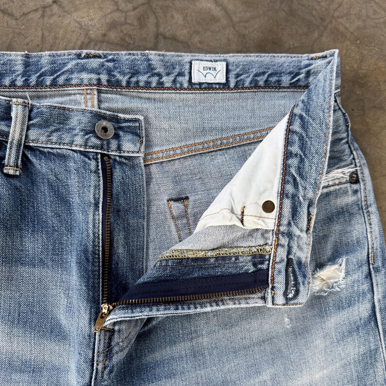 Pu&ograve; includere: Primo piano di jeans di denim blu chiaro con cerniera parzialmente aperta. I jeans hanno una chiusura con bottone e una piccola etichetta con il marchio "EDWIN". Il denim ha un aspetto sbiadito e usurato con alcuni dettagli strappati.