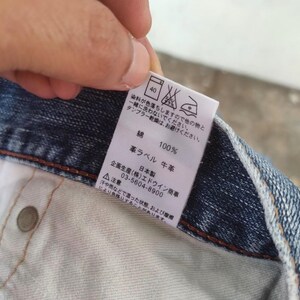 May include: Close-up of a denim garment with a white tag. The tag features Japanese text, care symbols, and the words "100%" and "leather label". The denim is a medium blue wash.