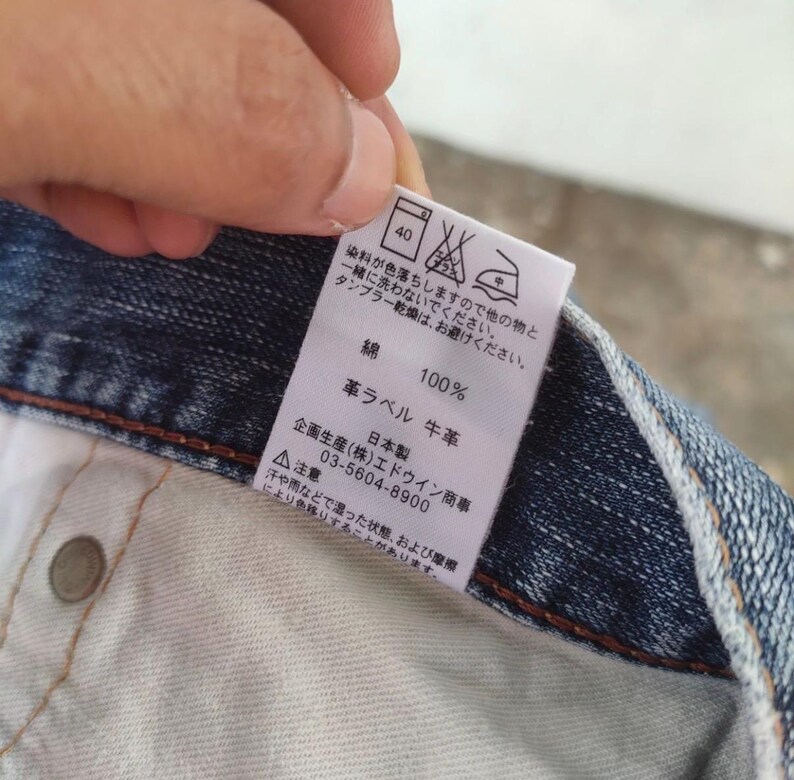 May include: Close-up of a denim garment with a white tag. The tag features Japanese text, care symbols, and the words "100%" and "leather label". The denim is a medium blue wash.