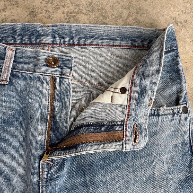 May include: Close-up of light blue denim jeans with a partially unzipped brass zipper. The jeans have a button closure and red stitching details. The fly is open, revealing the interior construction.
