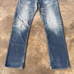 以下が含まれることがあります： 色あせた青色のデニムジーンズ。ダメージ加工が施されており、膝に破れがあります。ストレートレッグカットで、デニム全体に色あせや明るい部分があり、使い古された外観をしています。
