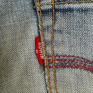 May include: Close-up of a section of blue denim jeans. A small red tag with the brand name "EDWIN" is sewn into the seam. The stitching is in orange and red thread, creating a textured pattern.