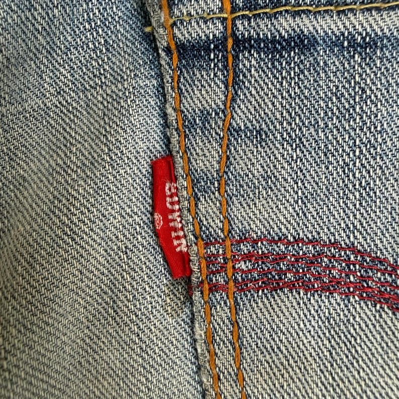 May include: Close-up of a section of blue denim jeans. A small red tag with the brand name "EDWIN" is sewn into the seam. The stitching is in orange and red thread, creating a textured pattern.