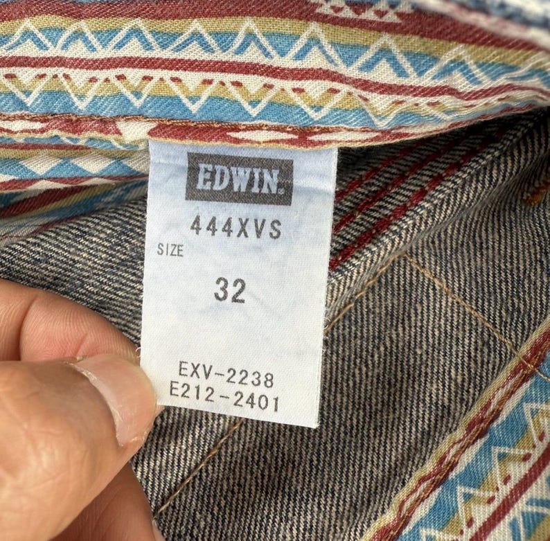 May include: Close-up of an Edwin jeans tag. The tag is white with black text, displaying the brand name, style number 444XVS, and size 32. The jeans have a patterned interior lining with blue, red, and tan geometric designs.