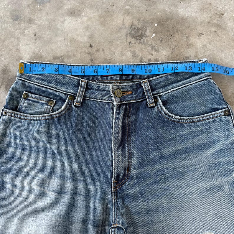May include: Close-up of blue denim jeans with a button closure and pockets. A blue measuring tape shows the waist size. The jeans have a classic five-pocket design and a faded wash.
