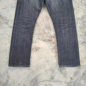 Pu&ograve; includere: Un paio di jeans in denim grigio scuro. I jeans hanno un taglio dritto e mostrano segni di usura. Il tessuto ha un aspetto strutturato, con sottili variazioni di colore. I jeans sono esposti su uno sfondo neutro.
