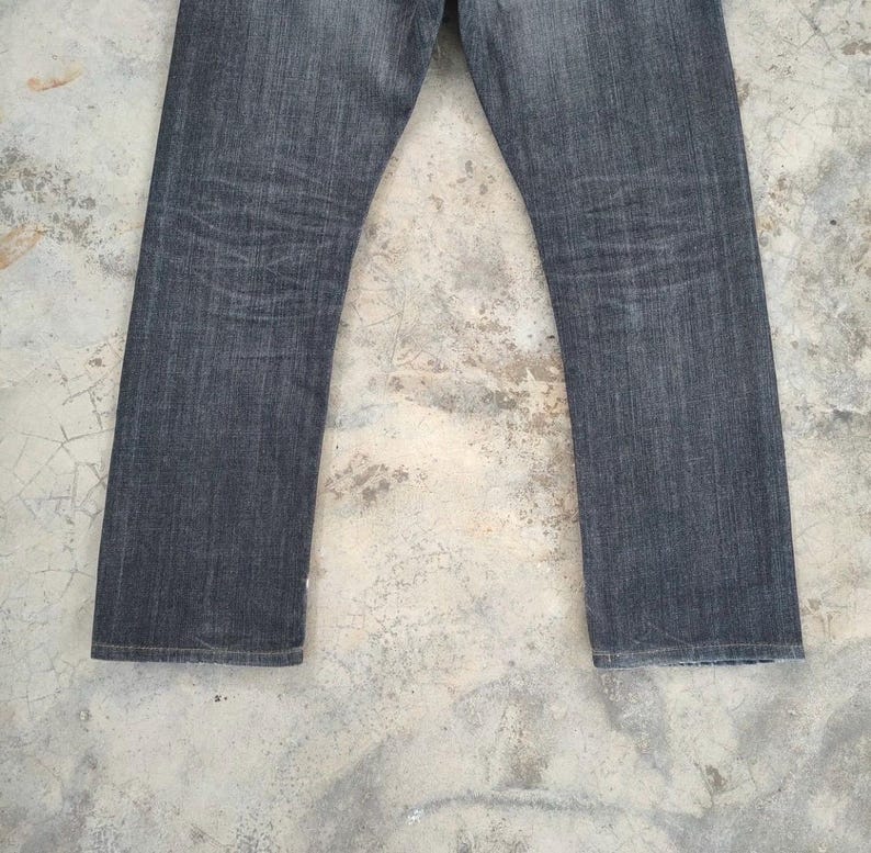 Pu&ograve; includere: Un paio di jeans in denim grigio scuro. I jeans hanno un taglio dritto e mostrano segni di usura. Il tessuto ha un aspetto strutturato, con sottili variazioni di colore. I jeans sono esposti su uno sfondo neutro.